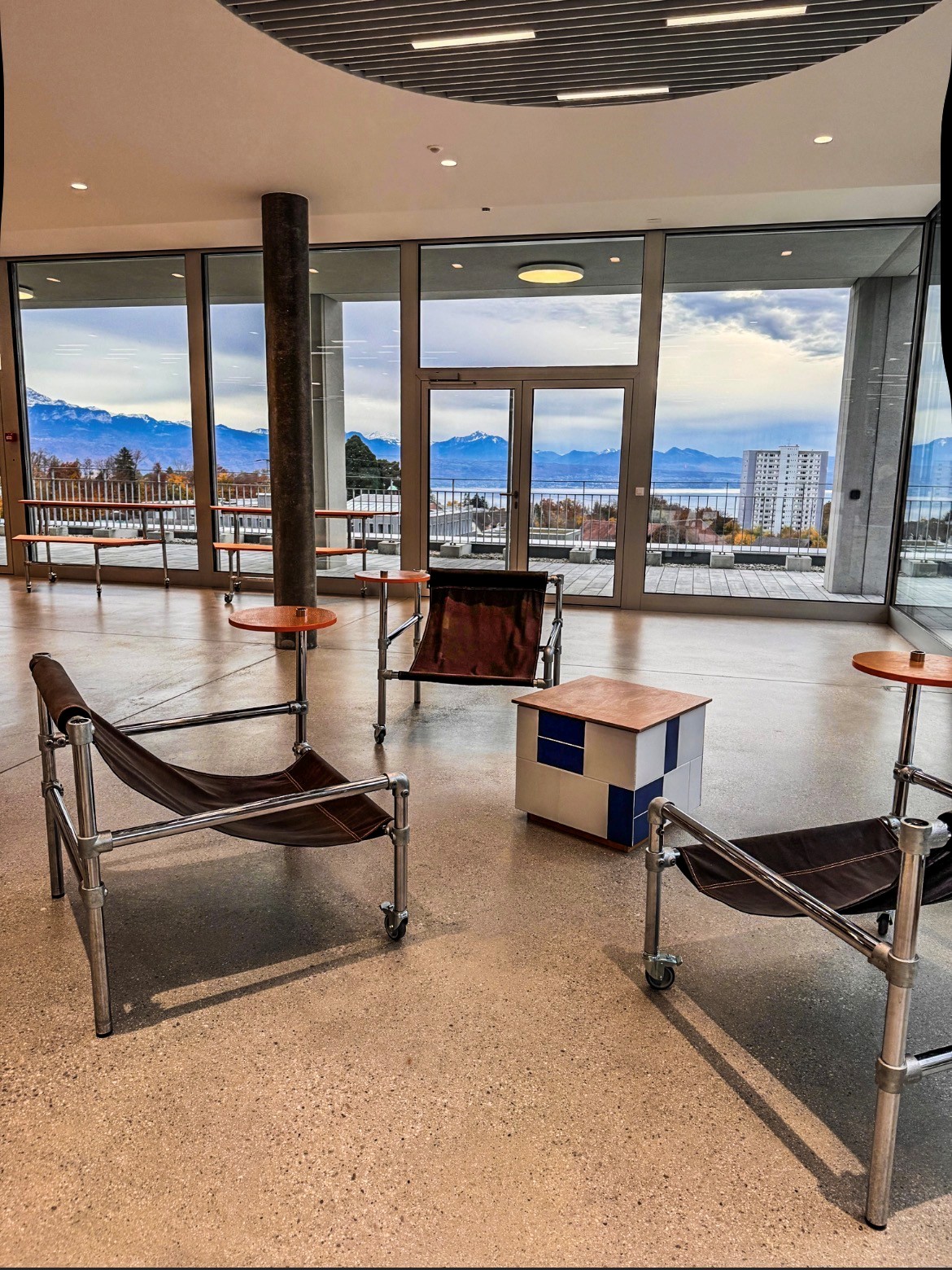 Salon moderne avec grandes baies vitrées donnant sur le lac, les montagnes enneigées et un ciel nuageux. Trois fauteuils en cuir marron à structure chromée sur roulettes et de petites tables orange sur sol en terrazzo clair.