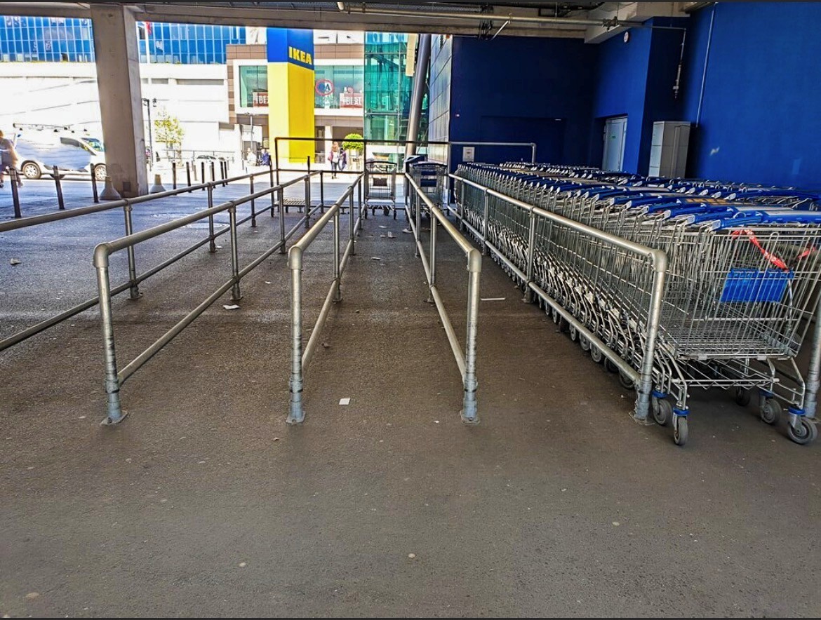 Zone d'accès d'un magasin IKEA : à droite, une longue rangée de caddies soigneusement empilés, dotés de poignées bleues. À gauche, des barrières métalliques mènent à l'entrée en traversant le sol gris. À l'arrière-plan, un grand logo IKEA jaune et bleu.