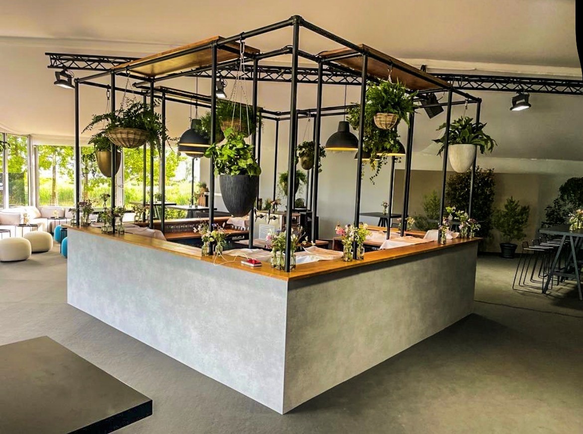 Comptoir moderne aspect béton gris et plateau bois dans une pièce lumineuse. Au-dessus, structure métallique noire avec plantes vertes suspendues et suspensions lumineuses. Petits arrangements floraux. Grandes fenêtres sur verdure.